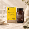 One Nutrition Methylated B-Complex food supplement shown with its yellow retail box and amber bottle, styled on a neutral fabric surface with a glass, ceramic bowl, and vase in the background.